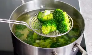 Frisch geernteter, wilder Brokkoli wird in einem Topf mit kochendem Wasser blanchiert. Ein Sieb hebt die leuchtend grünen Röschen hervor.