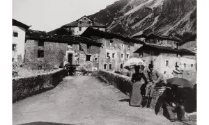 Un villaggio di montagna immortalato da Giovanni Verga fotografo. Coppie eleganti passeggiano con ombrelli, sullo sfondo case in pietra e pendii scoscesi.