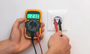 Un multimètre affiche 230V, tandis que ses sondes sont insérées dans une prise. La vérification de la prise terre est obligatoire pour la sécurité.