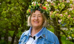 Glad kvinna med blomsterkrans, Bettan byvald, ler i solsken bland blommande träd.