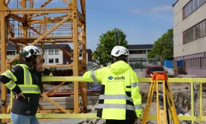 Två glada medarbetare i varselkläder och hjälmar på en byggarbetsplats. En stor kran syns i bakgrunden.