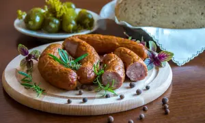 Kiełbasa podwawelska na drewnianej desce z rozmarynem i pieprzem. W tle ogórki i chleb.