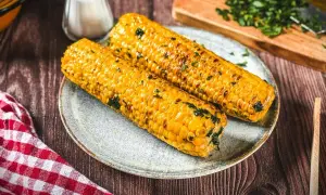 Zwei goldgelbe Maiskolben, perfekt gegrillt und mit Kräutern bestreut, liegen auf einem Teller. Ein Genuss für alle, die Maiskolben grillen lieben.