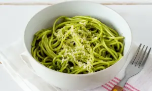 Miseczka spaghetti z zielonym, aromatycznym pesto z pokrzywy, posypana startym parmezanem. Obok leży widelec.