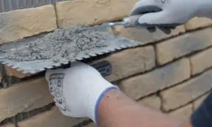 Ręce w rękawiczkach nakładają zaprawę na kamień dekoracyjny betonowy, przygotowując go do ułożenia na ścianie.