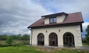 Nowy dom w Charsznicy z łukowymi oknami i brązowym dachem, otoczony zielenią i chmurami na niebie.