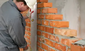 Murarz w kasku i rękawicach używa poziomicy do sprawdzania równości różnych rodzajów cegieł podczas budowy ściany.