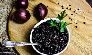 Schwarze, karamellisierte balsamico zwiebeln in einer Schüssel, garniert mit Petersilie. Daneben rote Zwiebeln und Pfefferkörner.