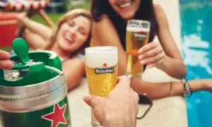 Un fusto di birra Heineken pronto per essere spillato, con due donne sorridenti che brindano con bicchieri di birra vicino a una piscina.