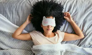 A woman with a large afro wears a cat-ear sleep mask, resting in bed. She's trying to figure out how long does it take to catch up on sleep.