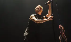 Lead singer of Nothing But Thieves, Conor Mason, belts into a microphone on a dark stage, his arm tattoos visible.
