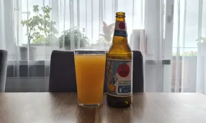 Szklanka z pomarańczowym napojem i butelka piwa bezalkoholowego mango ale na stole.