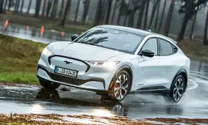 Biały Mustang Mach-E pokonuje kałuże na mokrej drodze, otoczony lasem.