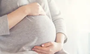 Brzuszek w 6 miesiącu ciąży, otulony szarym materiałem. Dłonie delikatnie go obejmują, czuć radość oczekiwania.