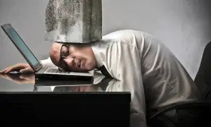 Man med sten på huvudet, utmattad vid sin laptop. Symboliserar digital stress och överväldigande arbetsbelastning.
