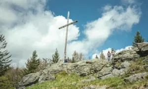 Wanderer auf einem felsigen Gipfel im Bayerischen Wald unter einem Holzkreuz. Ein typischer 8-Tausender-Ausblick.