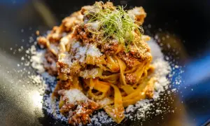 En portion pappardelle al cinghiale, toppad med riven ost och färska groddar, serverad på en svart tallrik.