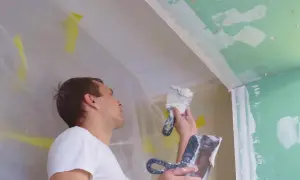 Mężczyzna wykonuje tynkowanie sufitu i narożników. Prace remontowe w toku, widoczne folie ochronne i narzędzia.