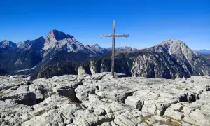 Holzkreuz auf felsigem Gipfel des Monte Piano, mit majestätischen Bergen im Hintergrund unter blauem Himmel.