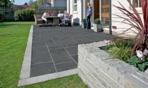Un cortile moderno con eleganti piastrelle per cortile esterno grigie, bordate da un acciottolato chiaro. Persone rilassate al tavolo, circondati da verde.