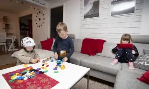 Tre barn leker med byggklossar och en surfplatta i ett vardagsrum. En pojke med hjälm bygger, en annan pojke leker med klossar, och en flicka tittar på en surfplatta. Leo Carmona barnen har kul.