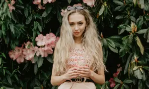 Młoda kobieta o blond włosach i słowiańskiej urodzie, w kwiecistej sukience, stoi na tle bujnej zieleni i różowych kwiatów.