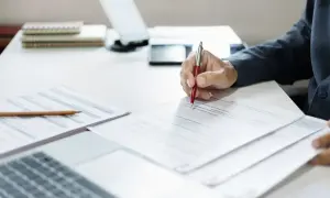 Ręka pisze długopisem po umowie. Czy na umowie zlecenie jest ubezpieczenie? Dokumenty i laptop na biurku.