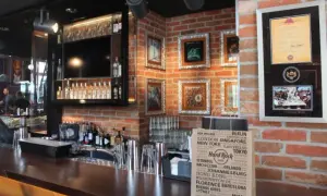 Wnętrze Hard Rock Cafe w Gdańsku: bar z butelkami alkoholu, ozdobne ramki na ścianie z cegły i torba z nazwami miast.