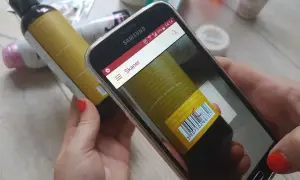 Ręce trzymają smartfon z otwartą aplikacją do sprawdzania składu kosmetyków. Na ekranie widoczny jest skaner kodu kreskowego skierowany na etykietę butelki.
