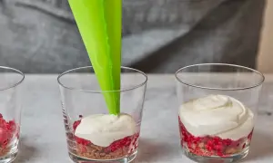 Cremige Masse wird mit Spritzbeutel in Gläser mit Himbeeren und Spekulatius-Bröseln gefüllt.
