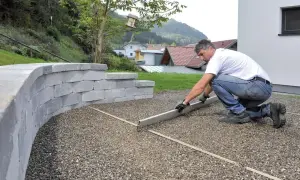 Mężczyzna wyrównuje podjazd z betonu szczotkowanego. Obok wznosi się mur z szarego kamienia.
