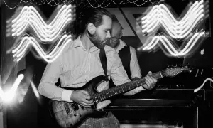 Muzyk gra na gitarze elektrycznej, a za nim widać drugiego członka zespołu. Efekt falujących świateł sugeruje kompensację ekspozycji.