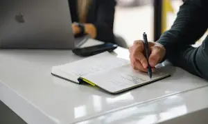 Dłoń pisząca długopisem w notatniku na białym biurku, obok laptopa.