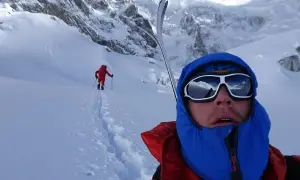Tom Ballard im Schnee, mit Skiern auf dem Rücken, blickt in die Ferne. Ein weiterer Skifahrer ist im Hintergrund zu sehen.