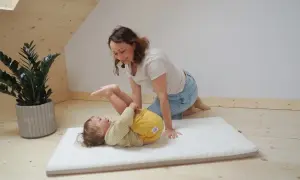 Mama bawi się z dzieckiem na miękkim materacyku do łóżeczka. Radosny maluch leży na plecach, a mama klęczy obok, uśmiechając się.
