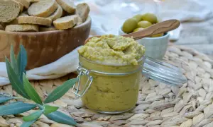 Un pot de tapenade verte, garni d'une cuillère en bois, est prêt à être dégusté avec des toasts et des olives.