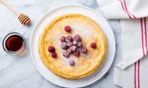 Ein Stapel goldbrauner Ofenpfannkuchen, garniert mit Himbeeren und Puderzucker. Daneben eine Schale Honig und ein Honiglöffel. Perfekt für den Thermomix!