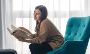 Kobieta z uśmiechem czyta książkę, wygodnie usadowiona w turkusowym fotelu do czytania.