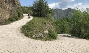 Ciclista su strada sterrata nel Parco Alto Garda Bresciano, tra rocce e vegetazione lussureggiante, con montagne sullo sfondo.