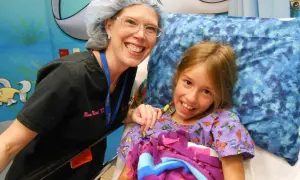 Niña sonriente en cama de hospital, junto a enfermera. Ella tiene síndrome de Marfan y recibe cuidados.
