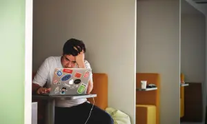 En frustrerad utvecklare funderar på hur mycket ska jag ta betalt per timme. Hans laptop är täckt av klistermärken.