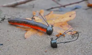 Zwei Angelköder, ein ned rig und ein weiterer, liegen auf nassem Sand neben einem Herbstblatt.