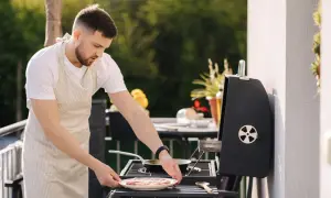 Mężczyzna w fartuchu przygotowuje mięso na grillu na balkonie. Czy można robić grilla na balkonie? Tak, jeśli jest to bezpieczne i zgodne z przepisami.