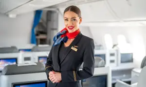 Uśmiechnięta członkini **cabin crew** w granatowym mundurze z czerwonym szalem, stoi na tle rzędów foteli w samolocie.