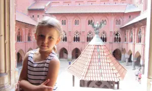 Dziewczynka w pasiastej sukience zwiedza zamek w Malborku. W tle widać ceglane mury i dach studni.