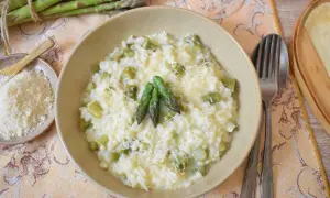 Kremowe risotto ze szparagami, posypane parmezanem, z kilkoma świeżymi szparagami na wierzchu. Obok miseczka z tartym serem.