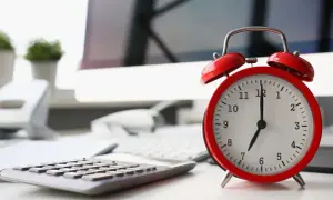 Sulla scrivania, un orologio rosso segna le 7. La calcolatrice e il computer suggeriscono che il giorno di riposo viene pagato, ma il lavoro attende.
