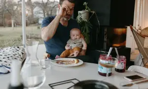 Trött pappa försöker äta frukost medan en bebis vaknar tidigt. Barnet sitter i pappans knä, en tallrik med mat står på bordet.