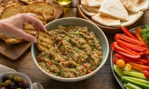 Une main trempe un pain grillé dans un bol de caviar d'aubergine, entouré de crudités, de pain pita et d'olives.