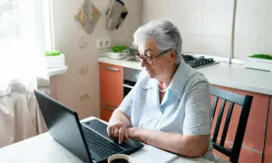 Starsza kobieta w okularach siedzi przy stole w kuchni i korzysta z laptopa.